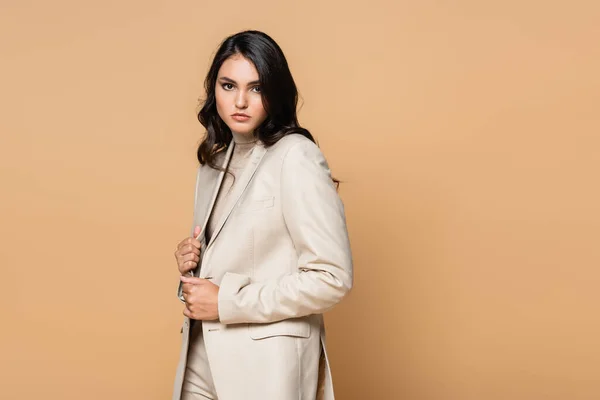 Brunette young model in trendy suit adjusting blazer isolated on beige — Stock Photo