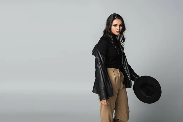 Young model in black leather jacket and turtleneck looking at camera while holding fedora hat on grey — Stock Photo