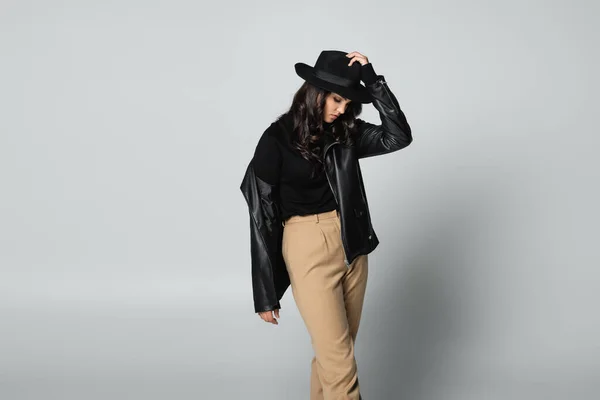 Молодая модель в черной кожаной куртке и водолазке, регулирующая шляпу Fedora на сером — стоковое фото