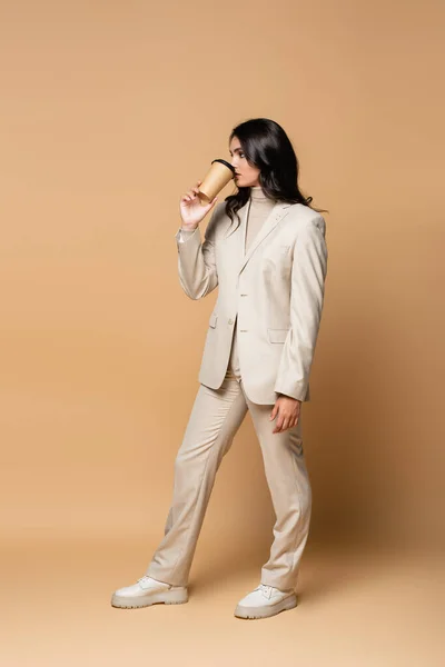 Full length of brunette young woman in trendy suit drinking coffee to go on beige — Stock Photo