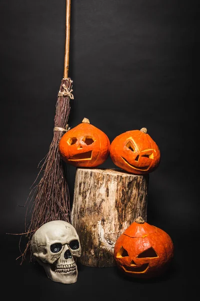 Vassoura perto de crânio e laranja esculpidas abóboras no toco de madeira no preto — Fotografia de Stock