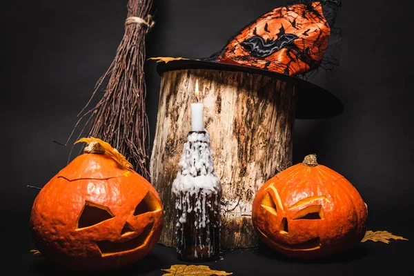 Candle with wax and orange carved pumpkins near witch hat on wooden stump and black background — Stock Photo