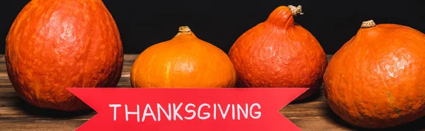 Ribbon with thanksgiving lettering near orange pumpkins on wooden surface isolated on black, banner — Stock Photo