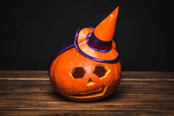 Creepy and carved pumpkin in small pointed hat on wooden surface isolated on black — Stock Photo