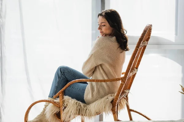 Mulher morena em camisola quente e jeans descansando em cadeira de balanço em casa — Fotografia de Stock