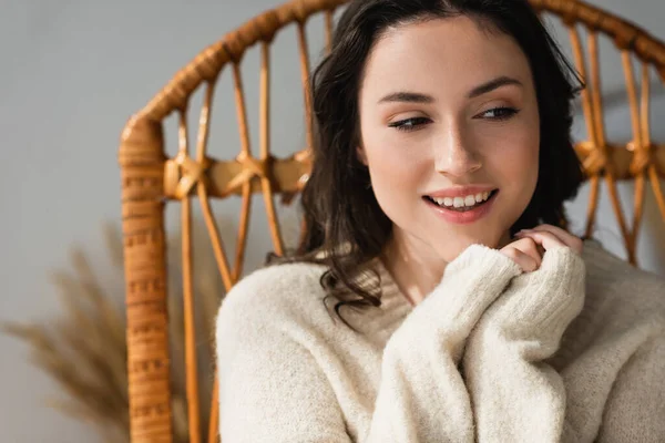 Mujer alegre mirando hacia otro lado mientras está sentado en silla de mimbre con las manos cerca de la barbilla - foto de stock