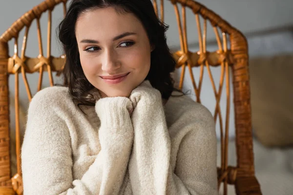 Mujer positiva en suéter caliente sentado en silla de mimbre y mirando hacia otro lado - foto de stock