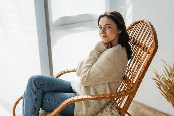 Young woman in warm sweater and jeans sitting in wicker chair and smiling at camera — Stock Photo