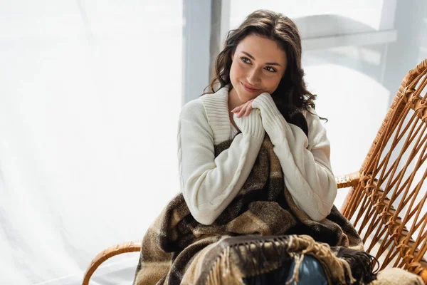 Joyful woman holding hands near face while sitting in wicker chair under soft blanket — Stock Photo