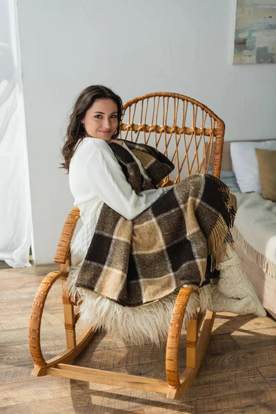 Zufriedene Frau blickt in die Kamera, während sie im Korbstuhl unter karierter Decke sitzt — Stockfoto