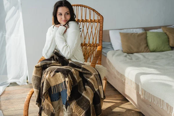 Femme souriante regardant loin tout en se reposant dans une chaise en osier sous une couverture à carreaux — Photo de stock