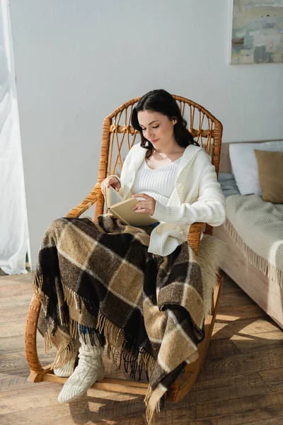 Donna sorridente seduta in sedia di vimini sotto coperta plaid e libro di lettura — Foto stock