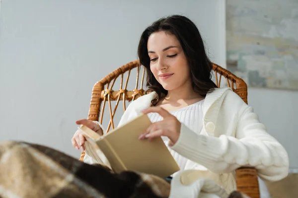 Jovem mulher sorrindo enquanto lendo livro na cadeira de vime sob cobertor quadriculado — Fotografia de Stock