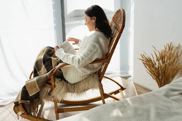 Vue latérale du livre de lecture femme brune dans un fauteuil à bascule sous une couverture à carreaux — Photo de stock