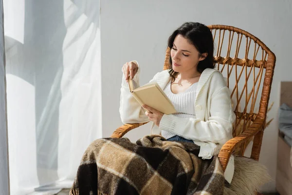 Brunette woman in white cardigan reading novel in whicker chair under warm blanket — Stock Photo