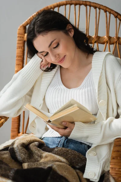 Счастливая молодая женщина сидит в плетеном кресле и читает роман — стоковое фото