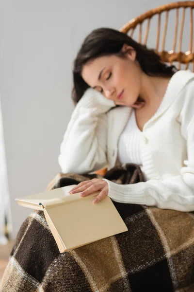 Donna sfocata dormire con libro in sedia di vimini sotto coperta plaid — Foto stock