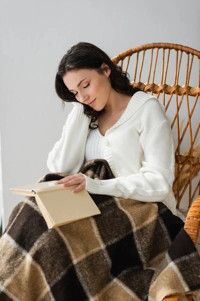 Молода жінка з книгою спить у плетеному кріслі під картатим ковдрою — стокове фото