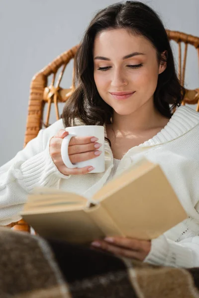 Glückliche Frau mit einer Tasse warmen Getränks, die zu Hause verschwommenes Buch liest — Stockfoto