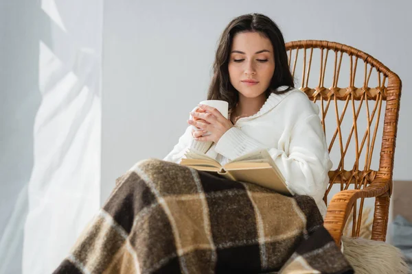 Брюнетка жінка сидить у плетеному кріслі з чашкою чаю і читає роман — стокове фото