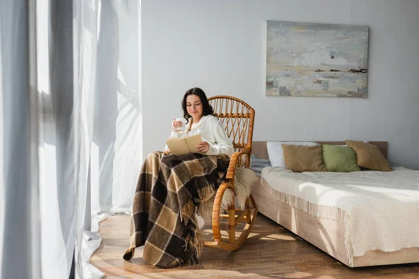 Brünette Frau liest Buch im Schlafzimmer, während sie im Schaukelstuhl unter karierter Decke sitzt — Stockfoto