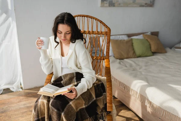 Жінка в білому кардигані читає книгу, сидячи в плетеному кріслі в спальні — стокове фото