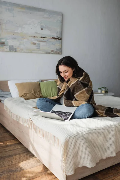 Lächelnde Frau sitzt auf dem Bett unter karierter Decke und schaut auf Laptop — Stockfoto