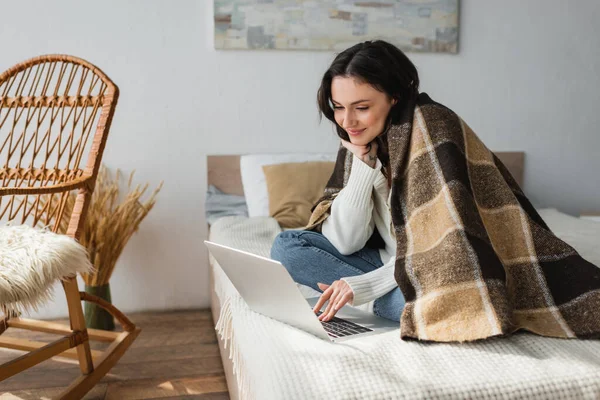 Lächelnde Frau mit Computer auf dem Bett, während sie unter karierter Decke sitzt — Stockfoto