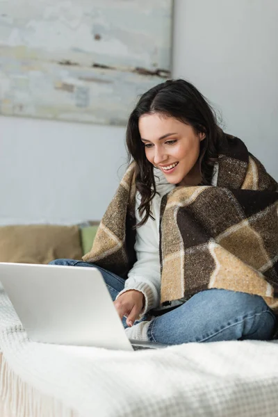 Happy young woman sitting in bedroom under plaid blanket and using laptop — Stock Photo