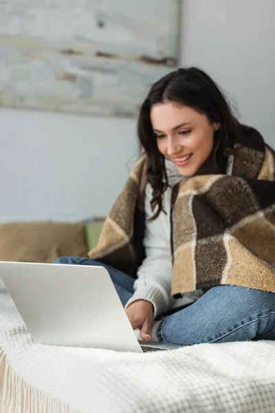 Blurred woman smiling while using laptop on bed under checkered blanket — Stock Photo