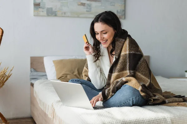 Довольная женщина сидит на кровати под теплым одеялом, держа кредитную карту рядом с ноутбуком — стоковое фото