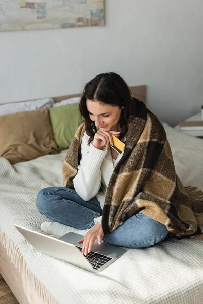 Mulher feliz com cartão de crédito usando laptop na cama sob cobertor quente — Fotografia de Stock