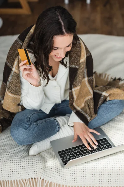 Glückliche Frau hält Kreditkarte in der Hand und tippt auf Laptop im Bett unter karierter Decke — Stockfoto