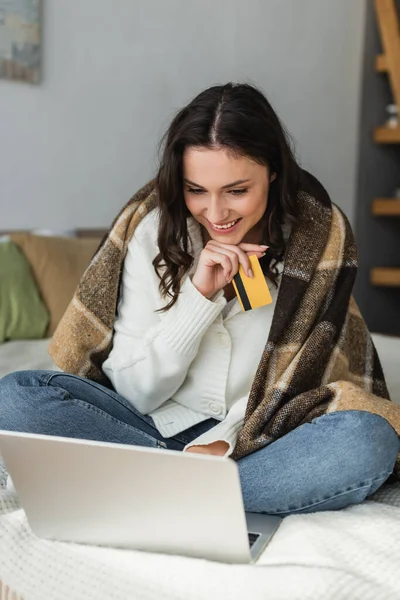 Femme gaie regardant ordinateur portable avec assis sous plaid couverture avec carte de crédit — Photo de stock