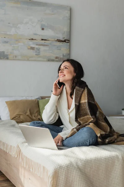 Радісна жінка говорить на смартфоні, сидячи на ліжку під ковдрою біля ноутбука — стокове фото