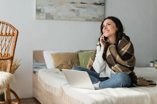Femme joyeuse assise sur le lit près d'un ordinateur portable et parlant sur un téléphone mobile — Photo de stock