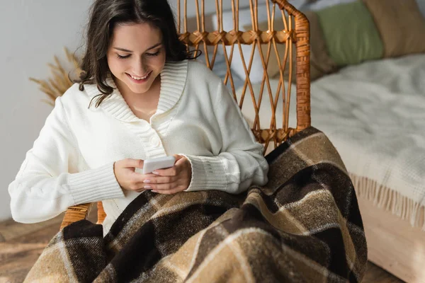 Cheerful woman sitting in wicker chair under checkered blanket and typing on mobile phone — Stock Photo