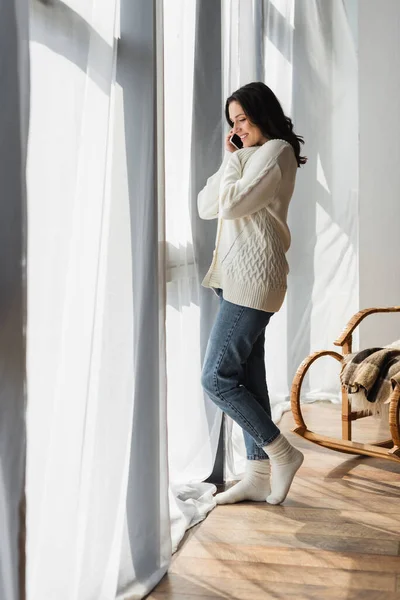Visão de comprimento total da mulher em cardigan de malha e meias quentes falando no celular perto da janela — Fotografia de Stock