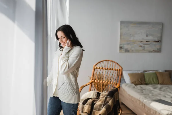 Улыбающаяся женщина разговаривает по смартфону, стоя у окна в спальне — стоковое фото