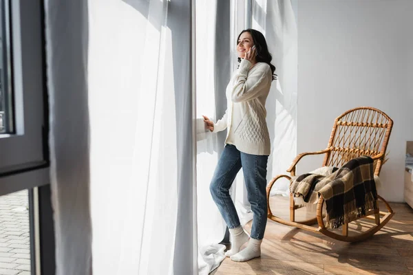 Full length view of woman in warm cardigan standing near window and talking on cellphone — Stock Photo