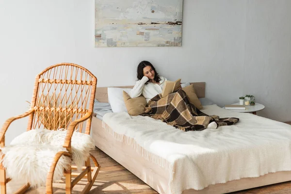 Brunette woman reading book in bed under plaid blanket near wicker chair — Stock Photo