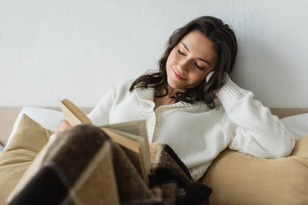 Mulher sorridente em cardigan branco livro de leitura na cama sob cobertor xadrez — Fotografia de Stock