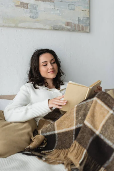 Jovem mulher positiva em cardigan branco livro de leitura na cama sob cobertor quente — Fotografia de Stock