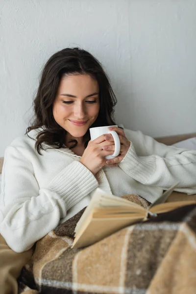 Glückliche Frau in warmer Strickjacke trinkt warmen Tee und liest Buch im Bett — Stockfoto