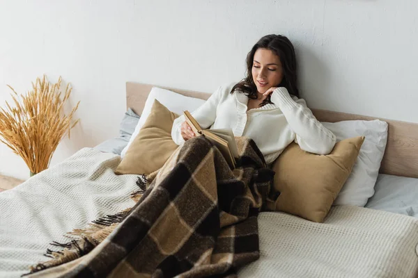 Lächelnde Frau liest Buch, während sie auf Kissen unter karierter Decke liegt — Stockfoto