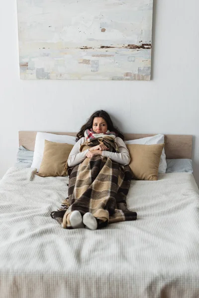 Visão comprimento total da mulher congelada em meias quentes deitado na cama sob manta xadrez — Fotografia de Stock