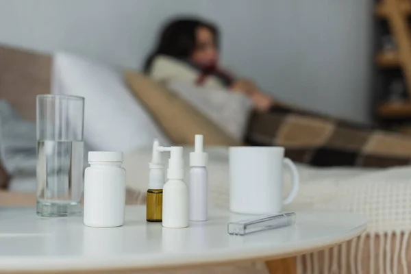Foco seletivo de recipientes com medicação, termômetro e bebidas na mesa de cabeceira perto de mulher doente em fundo turvo — Fotografia de Stock