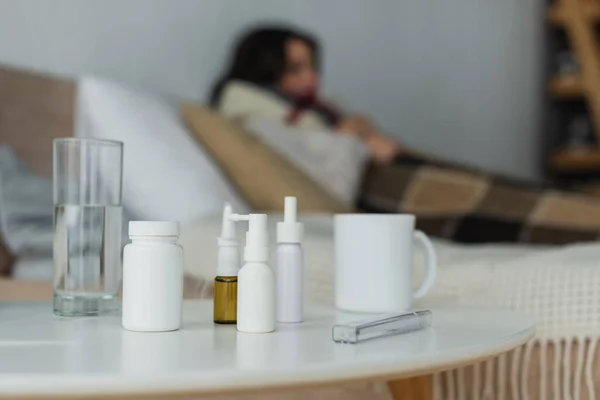 Selective focus of containers with medication, thermometer and drinks on bedside table near sick woman on blurred background — Stock Photo