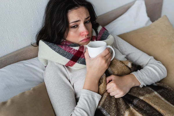 Malade et bouleversé femme couché sous couverture à carreaux avec tasse de boisson chaude — Photo de stock