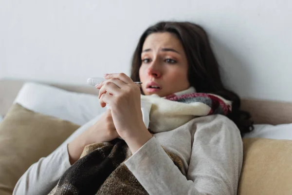 Donna scioccata guardando il termometro mentre giaceva a letto sotto la coperta plaid — Stock Photo